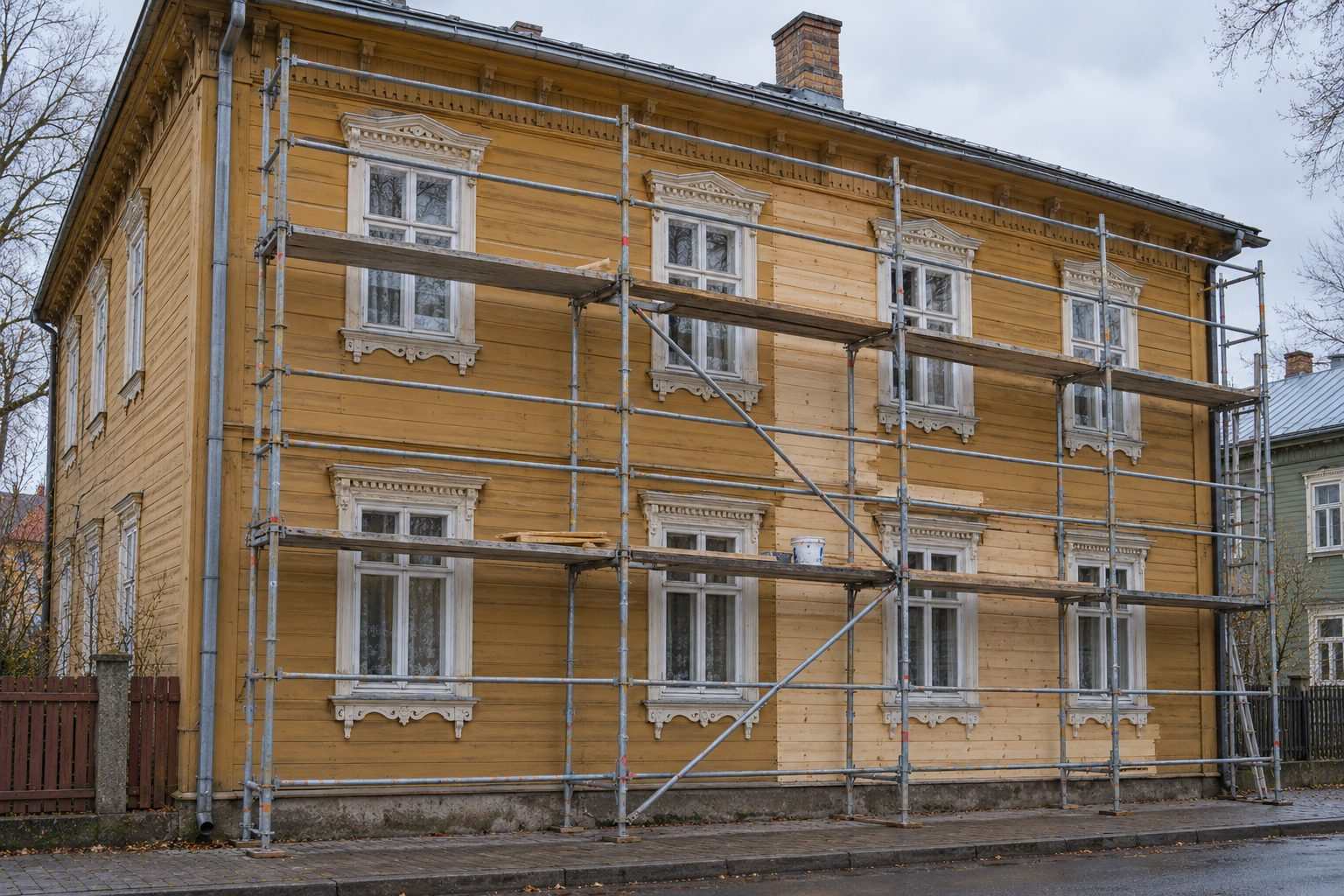 Fasādes sastatnes pie divstāvu koka mājas ar daļēji nomainītu apšuvumu